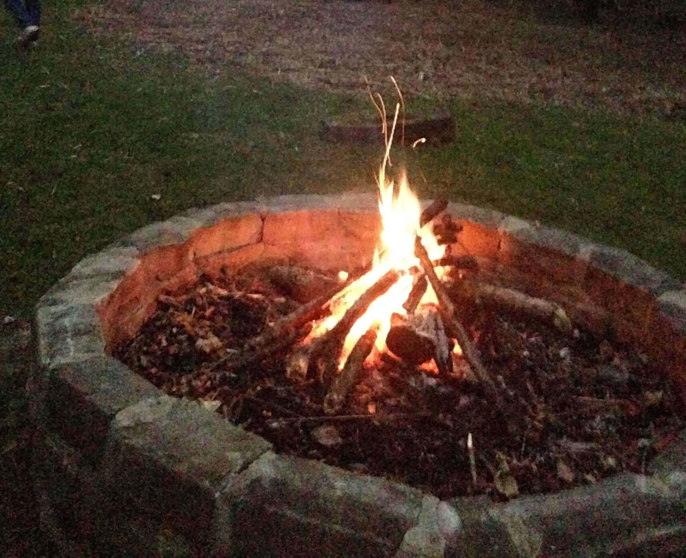 A campfire burning in a fire pit at dusk with people in the background.