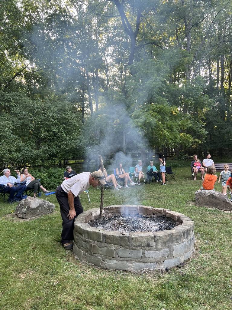 People gathered around a smoky outdoor fire pit in a wooded area.