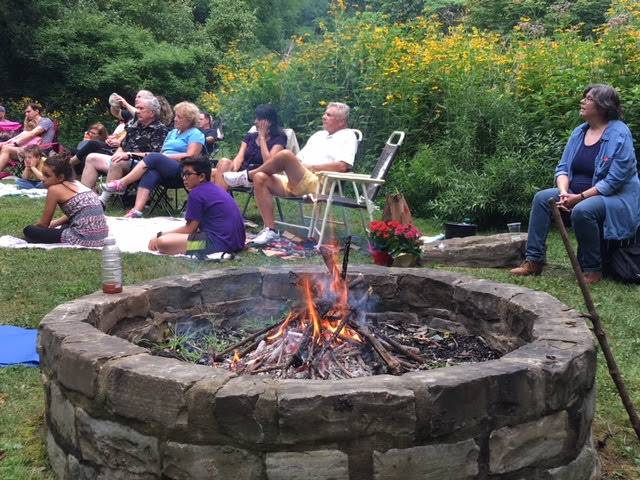 People gathered around a stone fire pit outdoors in a garden setting.