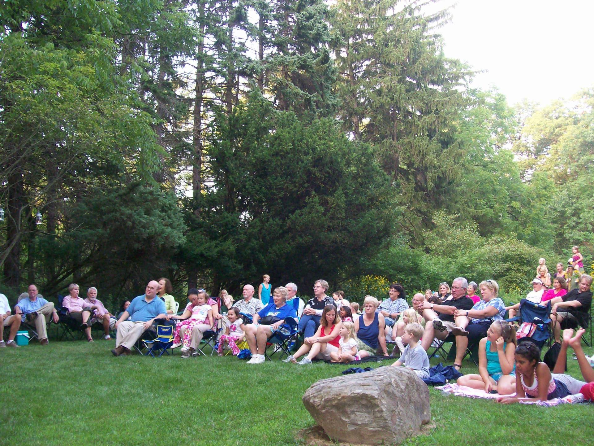 A large group of people gathered outdoors, sitting on grass and blankets.