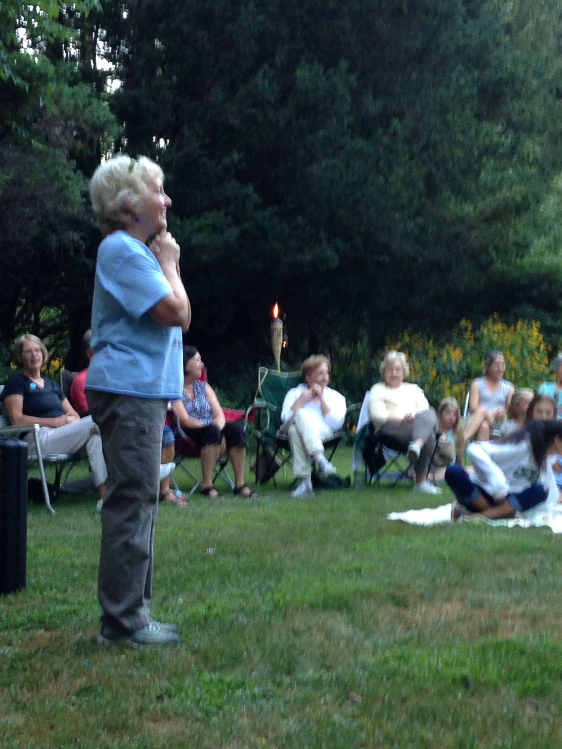 An elderly woman speaking outdoors to a seated audience in a garden.