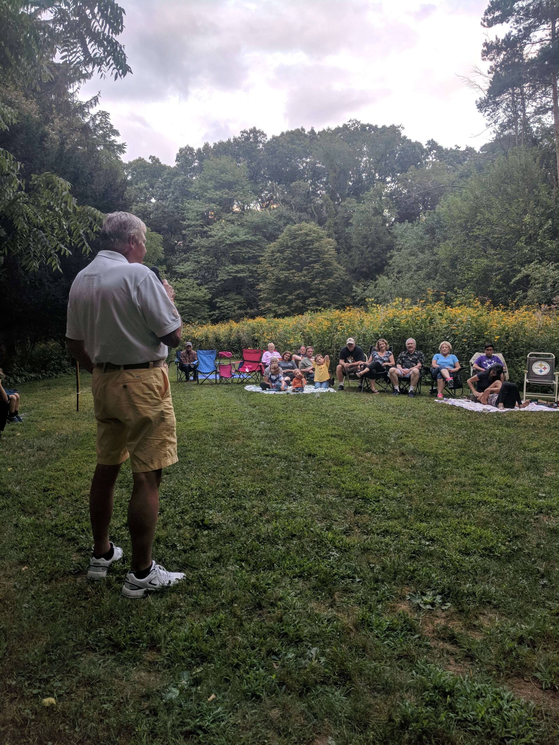 Man speaking to an outdoor audience in a garden setting.