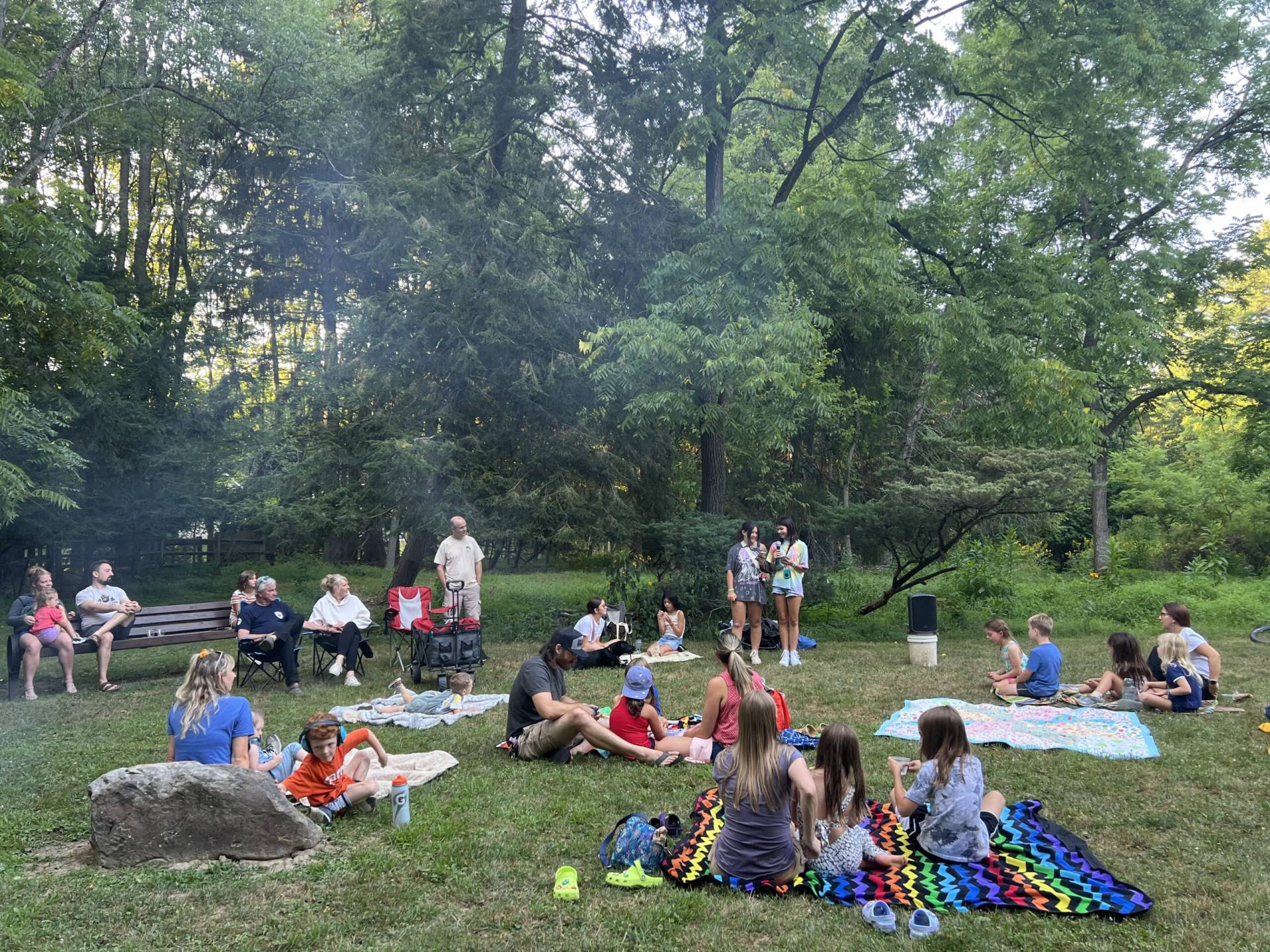 A diverse group of people gathered outdoors in a forest clearing, sitting on blankets and chairs.