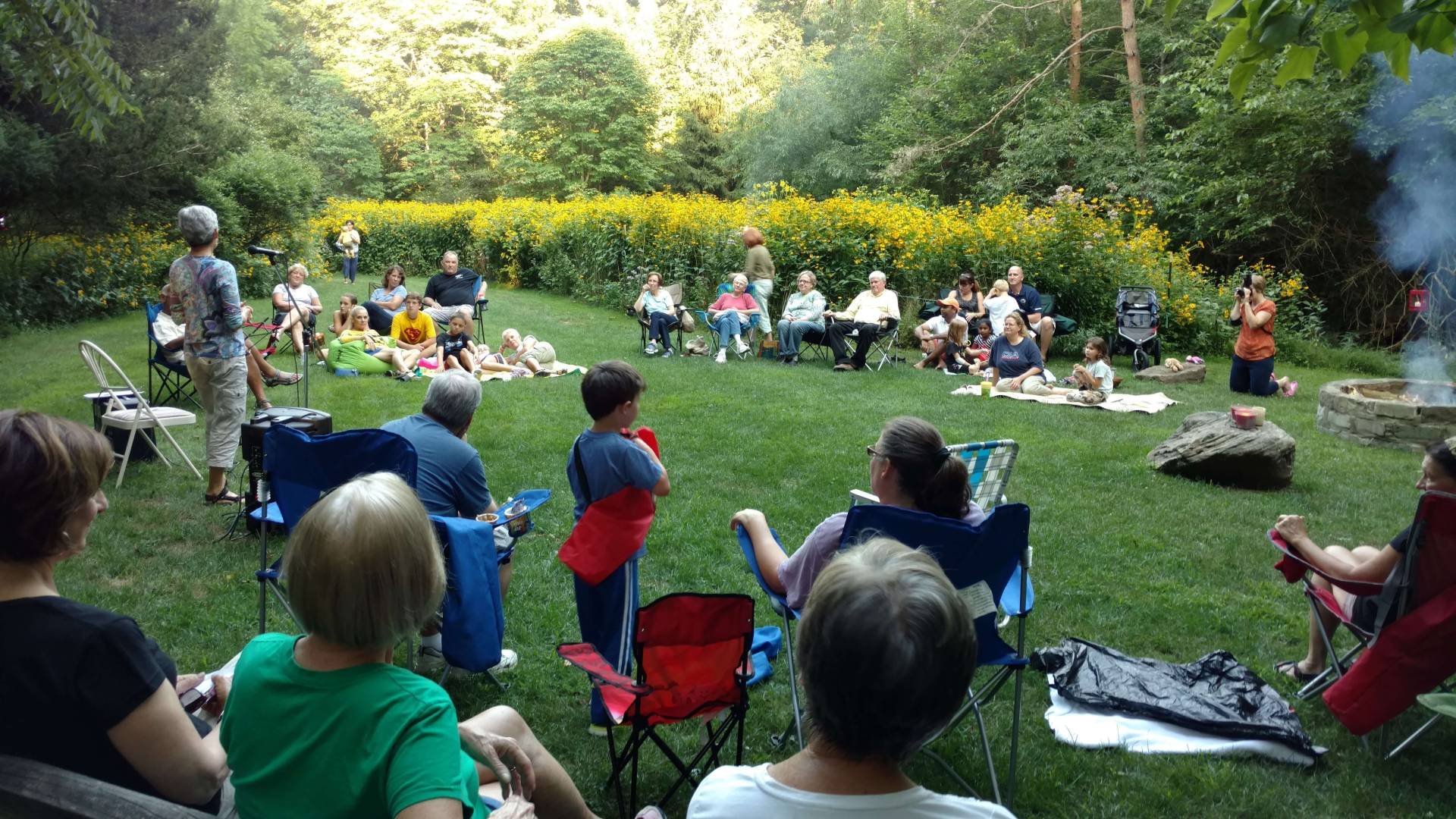People gathered outdoors sitting on grass and chairs in a park.