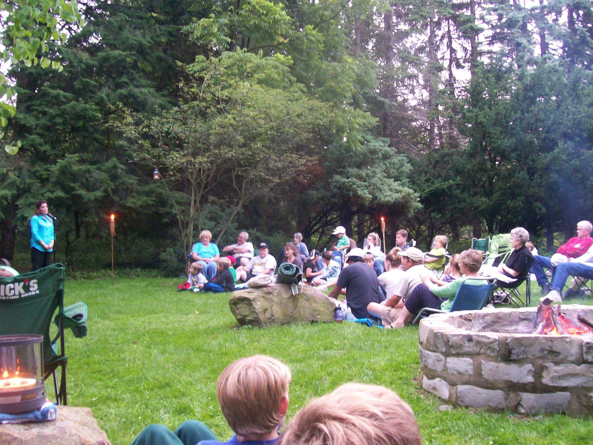 People gathered outdoors around a fire pit in a wooded area.