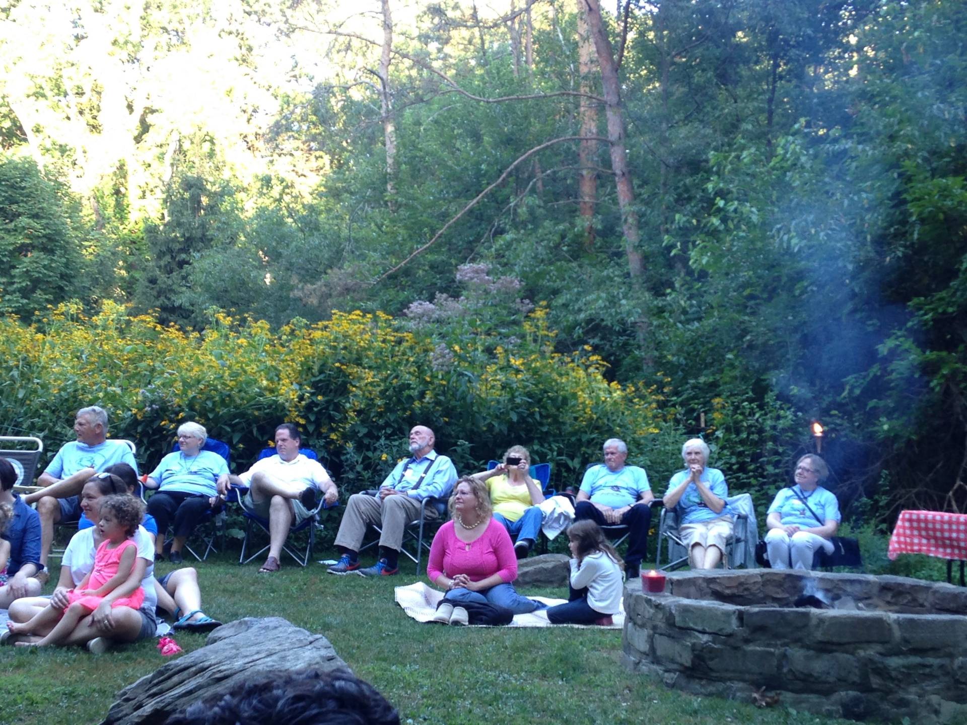 Group of people sitting outdoors near a campfire in a forest setting.