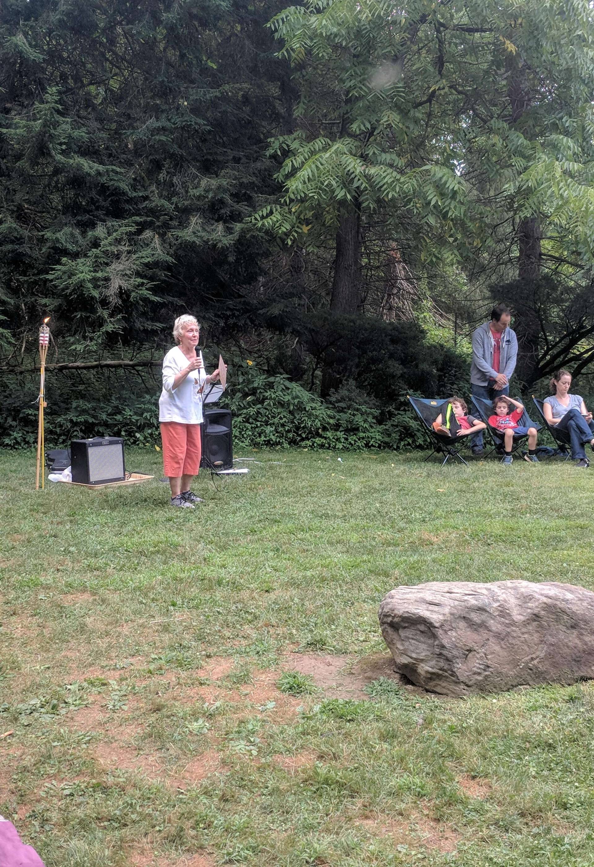 An elderly woman speaking outdoors with a small audience seated nearby.
